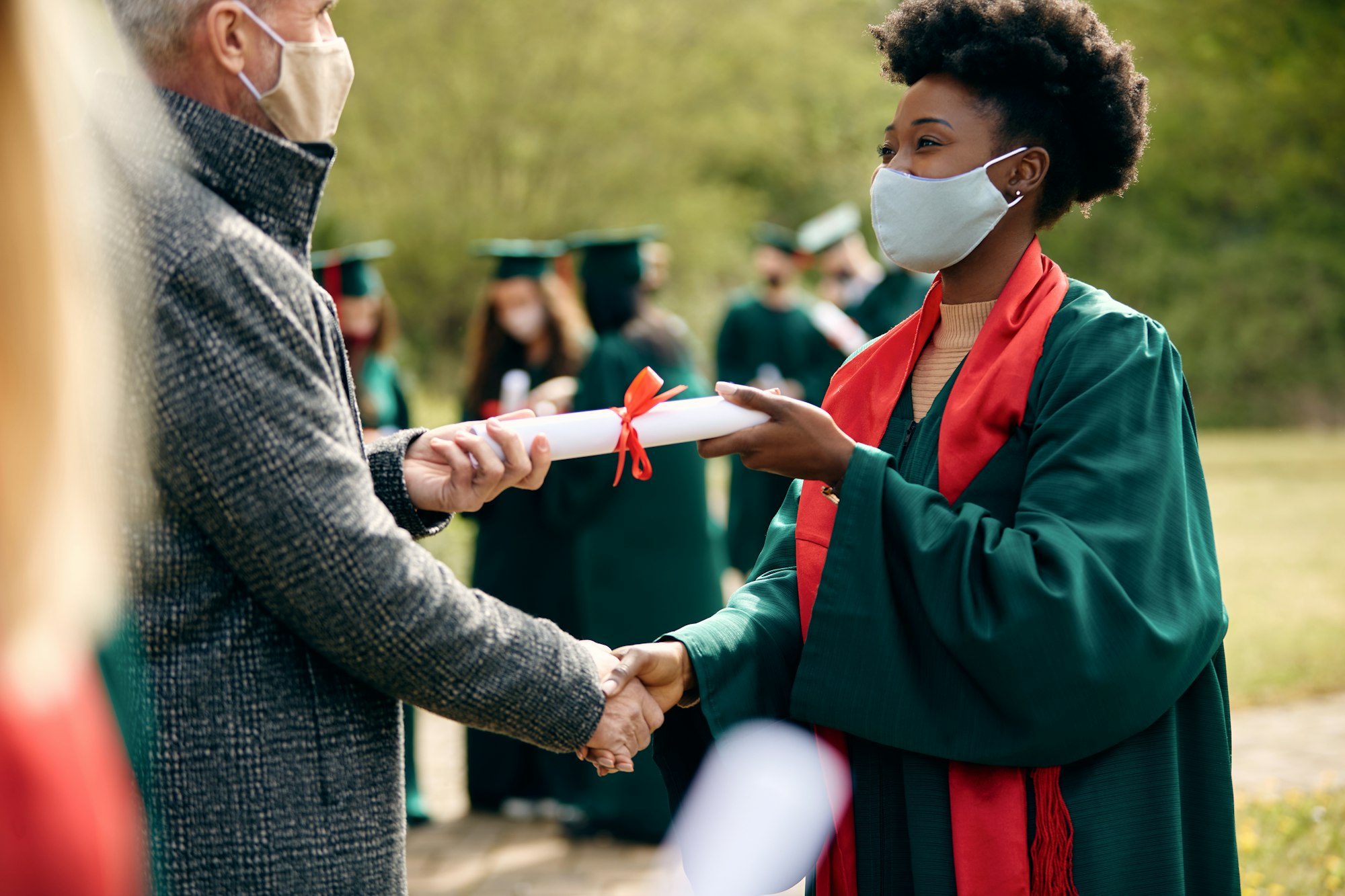 Happy Black student receiving diploma from professor on graduation day during coronavirus pandemic.